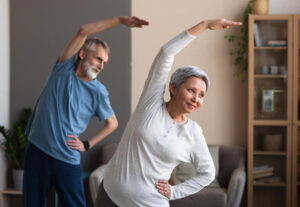 Two older adults in a exercise class. A man in a blue shirt in the back, and a women in a white shirt in the front.