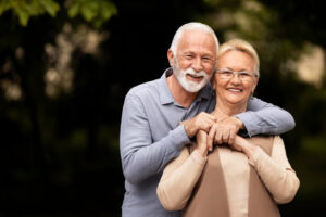 An older couple smiling outdoors. The older man has his arms around his wife's shoulders.