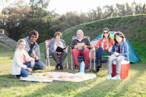 An older couple, and their family are having a picnic in the park. They are seated in lawn chairs next to their kids and grandkids.