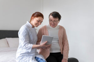 An older women with glasses in her room sitting next to her caregiver. They are looking at a mobile tablet.
