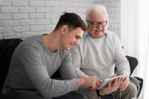 An older man and his son, watching something on a mobile tablet. The older man is wearing headphones.