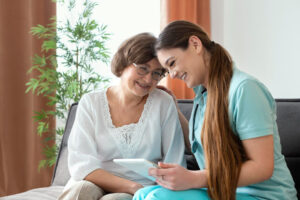 An older women sitting on a couch next to her caregiver. They are looking at a mobile tablet, and laughing.