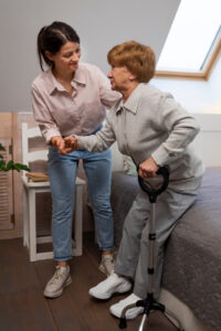 A caregiver helping an older women stand up from the side of her bed.