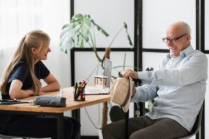 An older man and a caregiver are having a meeting in an office. The man is across the the caregiver, she is on the left, him on the right. They are laughing.