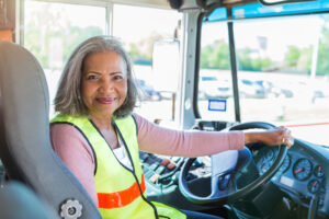A senior female bus driver turns to look at the camera from the driver's seat of her school bus. She has one hand on the steering wheel.