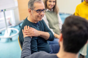 Senior man getting emotional support other pasticipant during group therapy session. Man putting hand on shoulder of upset senior male member and supporting him at community center.